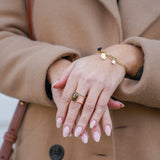 Zoom sur les mains d'une femme portant une bague en acier inoxydable avec une pierre en œil de tigre et un bracelet à breloques en nacre et noir, combinant élégance et style unique.